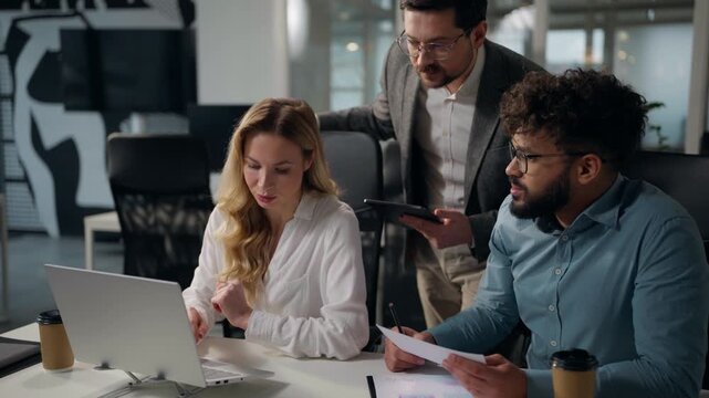 Three business people with document meeting with laptop talking multiethnic cooperation in global company multiracial colleagues at table analysing finance group office employees with computer working