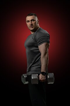 Strong athletic male bodybuilder holding heavy steel dumbbells while posing for a portrait in a dark studio setting with a dramatic red circular spotlight background.