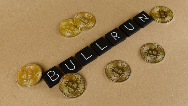 Black letter tiles spelling BULL RUN on a brown background, surrounded by shiny golden cryptocurrency coins, symbolizing market growth and financial trends