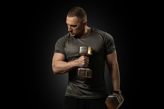 Focused, athletic male athlete lifts a dumbbell for bicep curl training inside a dark studio. High-contrast lighting highlights his muscular arm and physical fitness dedication.