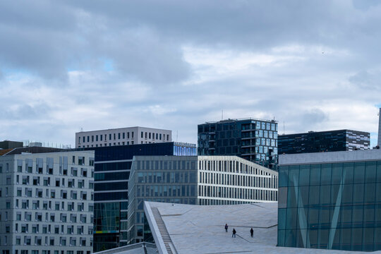 Modern architecture and office buildings shape the Oslo cityscape as pedestrians cross a sloping public space beneath a cloudy urban skyline