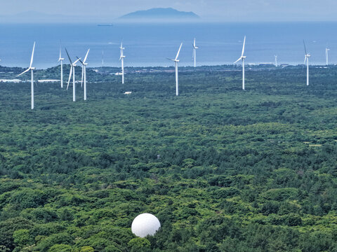 Aerial view of a wind farm with multiple turbines situated in a dense green forest near the coast, featuring a prominent white radome in the foreground, South Korea.