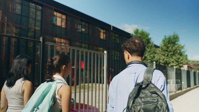 Three high school students walk together towards the school building carrying their backpacks. Friends are seen heading to campus on a bright, sunny day, ready for learning.