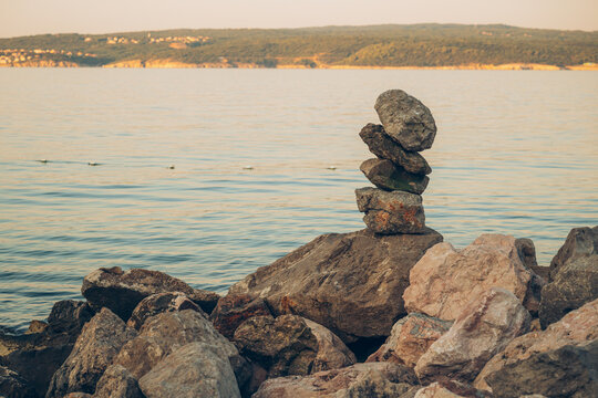 Balanced stack of stones on a rocky shore overlooking calm sea and a distant coastline at golden hour, evoking serenity, balance, meditation, and coastal landscape tranquility.
