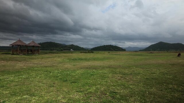 Slow pan across a vast green grass field with thatched bamboo pavilions below green mountains and dramatic monsoon storm clouds
