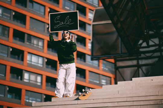Young woman holding a sale sign with excitement, standing on steps in front of modern architecture, featuring a skateboard as a symbol of youth culture and street style