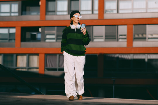 Stylish person wearing casual streetwear, white sunglasses, and headphones, checking a smartphone while walking outdoors in a modern urban environment with a building in the background