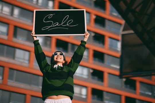 Happy young woman wearing sunglasses and a green striped sweater holding a sale chalkboard sign high above her head, celebrating a discount promotion outdoors in a city setting