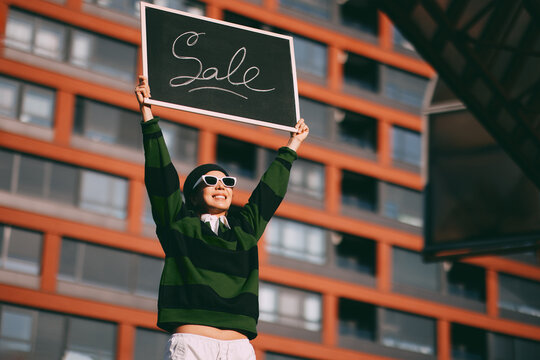 Young woman smiling and holding a chalkboard sign with the word sale written on it, celebrating shopping discounts and promotions in an urban environment