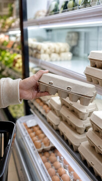 Close-up of a shopper holding a recycled paper egg carton beside refrigerated shelves stocked with eggs in a modern grocery store.