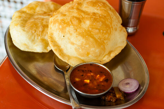 A plate of Chola bhatura, an Indian dish placed on a table.  Two deep-fried leavened breads a curry, along with some salads, are on the plate. This is the most common food eaten fo breakfast in India.
