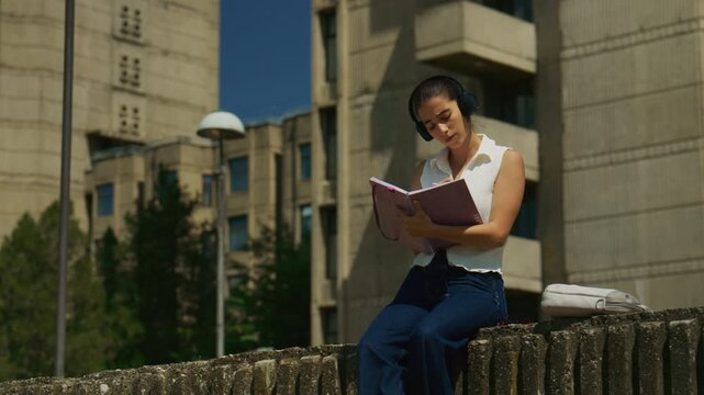 A high school student studies outdoors, wearing headphones and writing in a notebook. She is thoughtfully engaged in her academic work.