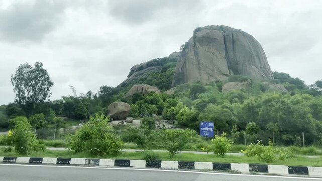 Full HD travel footage of a massive rocky hill beside a highway, with lush green vegetation, roadside trees and cloudy monsoon sky, capturing scenic road trip, nature landscape and rural tourism.
