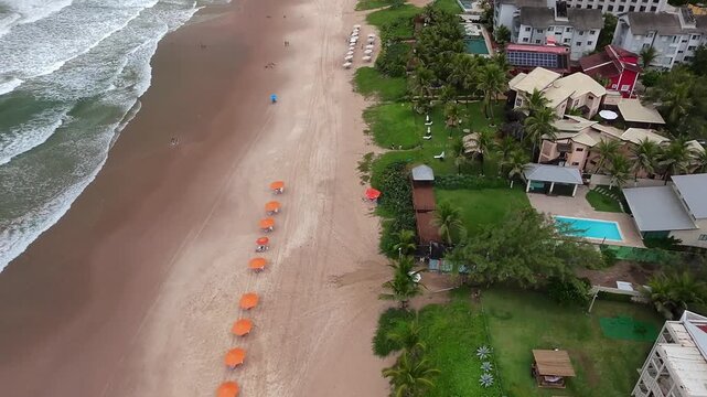 Aerial view of the sandy coastline of Maragogi featuring rows of orange umbrellas, coastal resorts with swimming pools, and lush palm trees in Maragogi, Alagoas, Brazil.
