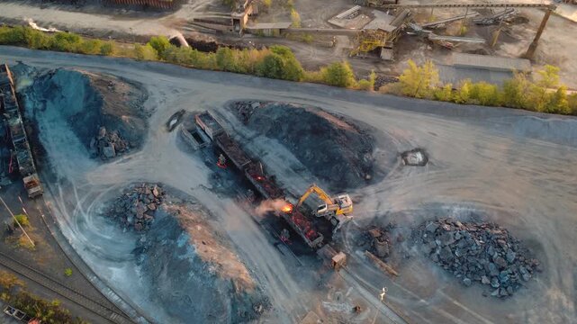 Top down aerial view of an excavator breaking up glowing hot slag in railway ladle cars at a steelworks, part of the slag processing in steel production