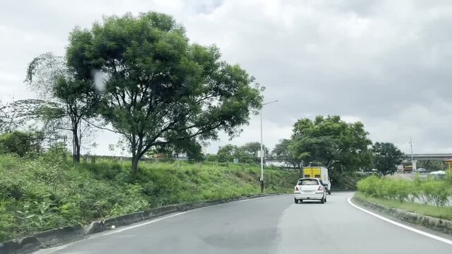 Full HD driving footage on a wet curving highway with cars and yellow truck passing through lush green roadside trees, cloudy sky and rural landscape, capturing monsoon road trip and travel journey.