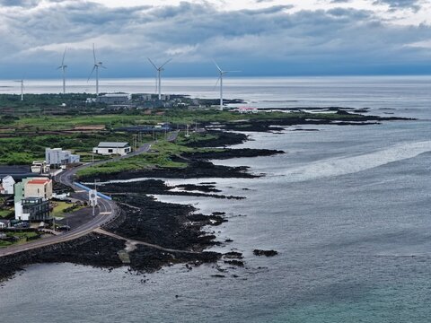 Aerial view of wind turbines along the rugged volcanic coastline with a coastal road and buildings in South Korea.