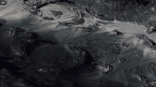 The surface of a planet with various circular craters and ridges. The terrain is dark with sharp contrasts and deep shadows. Details suggest geological features on a distant world.