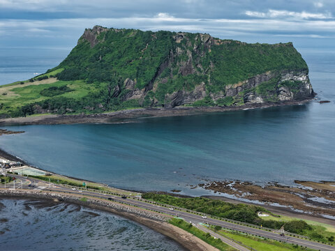 Aerial view of Seongsan Ilchulbong volcanic tuff cone rising above the coastal road and blue waters of Jeju Island, South Korea.