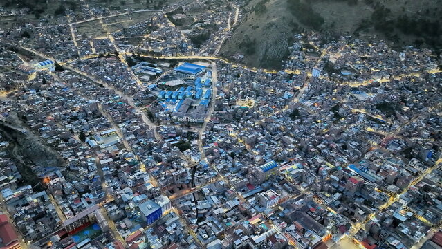Aerial view of the dense urban landscape of Comas at dusk, with illuminated streets winding through residential buildings and hillsides in the background, Comas, Provincia de Lima, Peru.