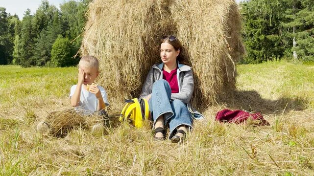 A mother and young boy sit by a haystack in a sunny meadow. The happy child eats a snack playfully while she relaxes. They feel joyful and peaceful during their calm outdoor break.