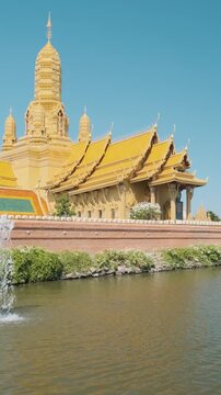 Ornate golden Thai temple complex with spire and traditional roof beside a pond with fountain in Ancient City, Bangkok, Thailand. Cultural heritage, traditional architecture. Travel destination