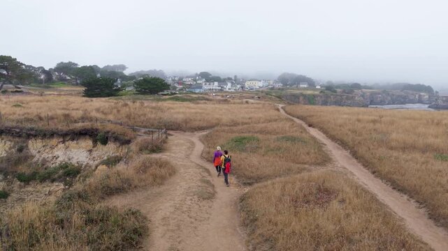Two people hike on the coastal trail in Mendocino, California as fog surrounds the area and ocean waves crash on cliffs below.