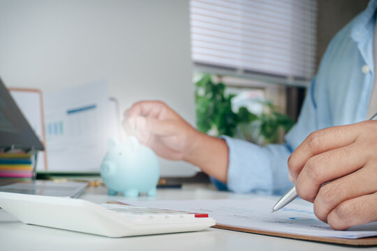 Personal Financial Concept, Person writing notes on financial report document with calculator and background blue piggy bank, personal budgeting, money saving, Digital and paper wealth management.