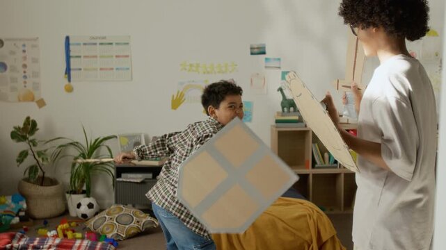 African American kid and teenager play-fighting with homemade cardboard swords and shields in bedroom. Video suitable for family lifestyle campaigns or creative play promotions