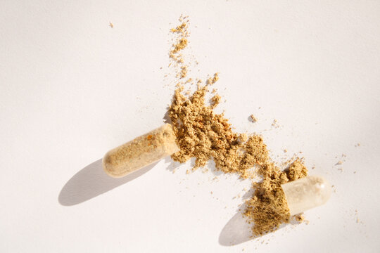 Top view of an open translucent capsule spilling natural herbal powder onto a clean white surface with bright lighting and distinct shadows.