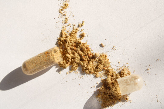 Herbal dietary supplement capsule opened with brown powder spilled on a clean white surface in bright direct sunlight with sharp shadows.