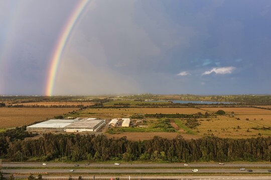 Aerial view of the landscape featuring a highway, agricultural fields, and a vibrant rainbow stretching across the sky in Chinderah, New South Wales, Australia.