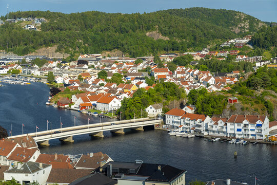 View of the coastal town of Mandal with its characteristic white houses, red roofs, and a bridge spanning the river, surrounded by lush green hills, Mandal, Agder, Norway.