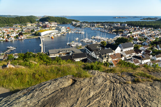 View of the coastal town of Mandal with its harbor, numerous boats, white houses, and a modern building connected by a bridge, seen from a rocky vantage point in Mandal, Agder, Norway.