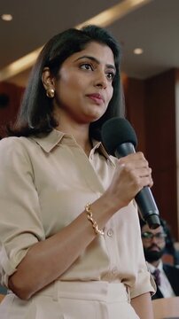 Vertical video: Standing Indian woman in tan blouse raising hand taking mic asking question in hall