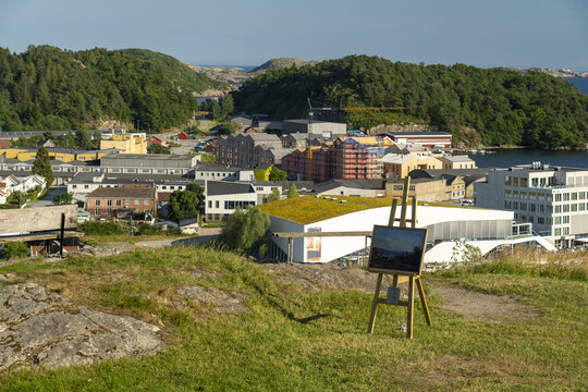View of the town of Mandal with an easel in the foreground, featuring various buildings, construction sites, and lush green hills in Mandal, Agder, Norway.