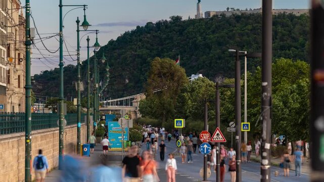 Budapest embankment promenade time lapse with passing pedestrians facing Gellert Hill and Liberty Statue in warm sunset light