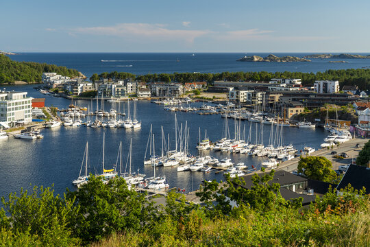 View of the marina in Mandal with numerous sailboats docked at the pier, surrounded by modern residential buildings and lush green trees in Mandal, Agder, Norway.
