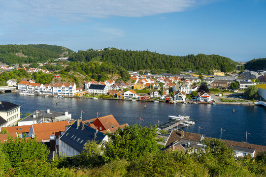 View of the historic town of Mandal with its characteristic white wooden houses, red-tiled roofs, and a calm river under a clear blue sky in Mandal, Agder, Norway.