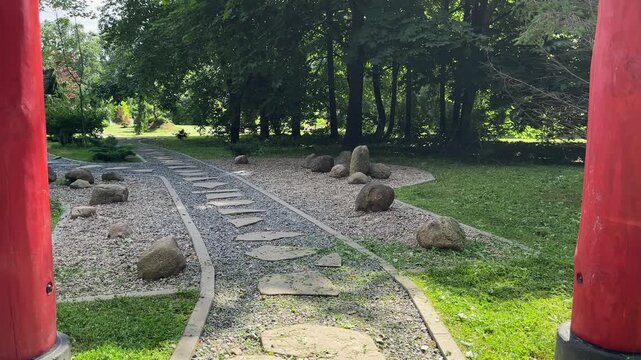 Traditional red torii gates border stone path in Japanese garden. Cobblestone walkway leads toward oriental pagoda house surrounded by trees. Perspective view frames Asian landscape