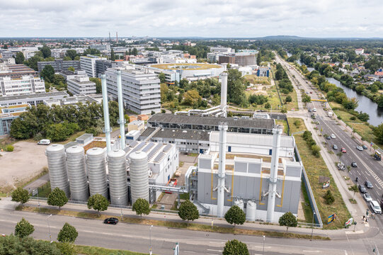 Aerial view of the BTB Heizkraftwerk Adlershof power plant with silver storage tanks and tall chimneys, surrounded by modern buildings, a road, and a canal in Berlin, Berlin, Germany.