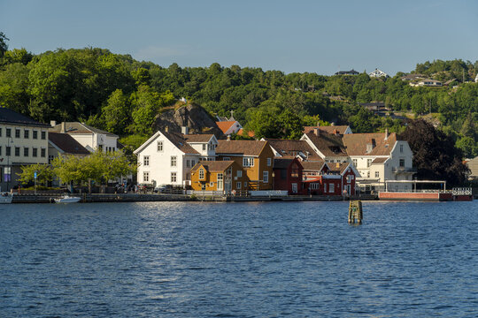 View of colorful wooden houses along the waterfront in the historic town of Mandal, with a lush green hillside in the background under a clear blue sky in Mandal, Agder, Norway.