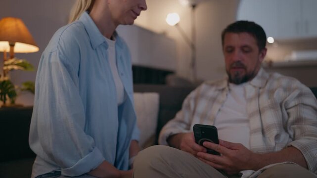 Woman gently touching her partner's leg while trying to talk, as he remains focused on his smartphone, illustrating emotional distance, communication problems and relationship difficulties.