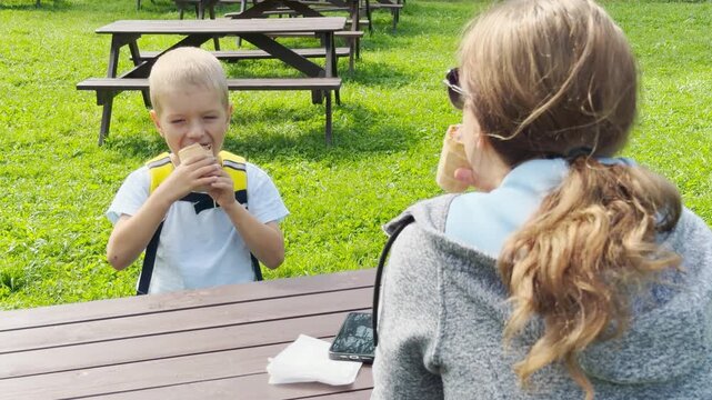 A mother and son sit at a wooden table and eat ice cream in a green park. They feel happy, joyful, and peaceful during this warm outdoor treat.