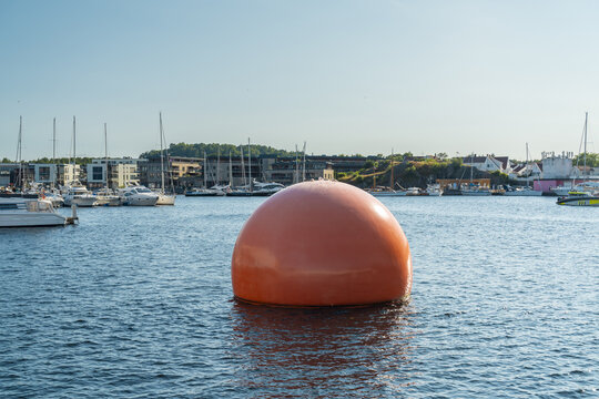 View of a large orange mooring buoy floating in the harbor with yachts and modern buildings in the background in Mandal, Agder, Norway.