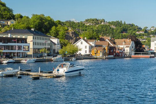 View of the waterfront in Mandal with moored motorboats, traditional white wooden houses, and lush green hills under a clear blue sky in Mandal, Agder, Norway.