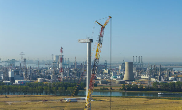 Aerial view of a wind turbine under construction with a large crane, set against an industrial complex with cooling towers and refinery infrastructure near a river under a clear blue sky.