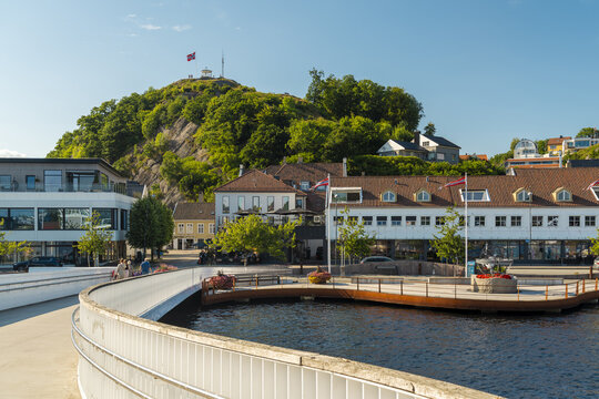 View of the waterfront area featuring a modern pedestrian bridge, a wooden deck, and the prominent Uranienborg hill in the background in Mandal, Agder, Norway.