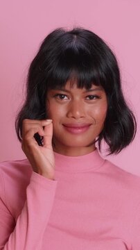 Beautiful Indonesian girl performs mouth zipping motion using fingers to show secret keeping act. Young female with black bob hairstyle smiles after locking lips against pink studio backdrop.