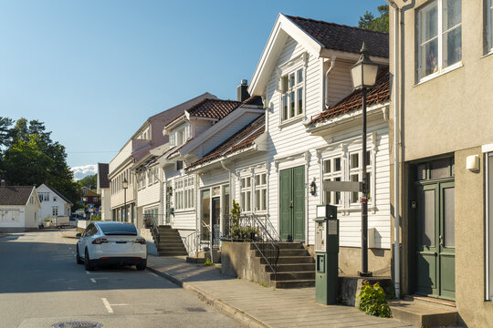 View of a quiet street with traditional white wooden houses, a parked white electric car, and a vintage street lamp in Mandal, Agder, Norway.